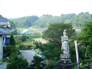 住宅や田畑が広がる風景を見つめている緑に囲まれた高台に建つ観音像の後ろ姿の写真