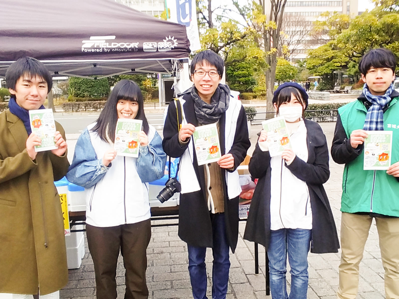 屋外のテントの前で、五人の若者が並んで立ち、それぞれチラシを手に笑顔でカメラに見せている様子の写真