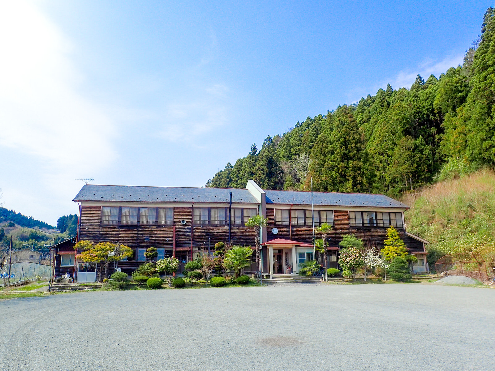 広い砂利の敷地に廃校となった木造二階建て校舎があり、周りに山や森があるさんさん館の外観写真