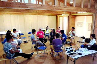 十数人の参加者が木造の室内で円く座り、中央に座ったピンク色のシャツを着た男性が話をする様子を少し上から撮影した写真