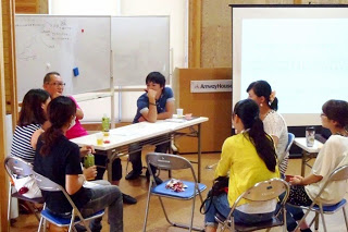 資料が映し出されたスクリーンの前で、7人の参加者が白いテーブルを囲んで活発に意見を交わしている写真