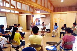 木造の明るい室内で、円になって椅子に座った参加者たちが紙を見ながら意見を交換している様子の写真