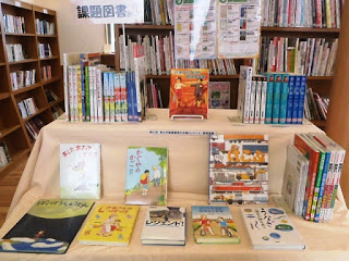 図書館の本棚に子供向けの絵本や読み物が並べられた展示コーナーがあり、中央に「課題図書」と書かれた看板が立っている課題図書コーナーの写真