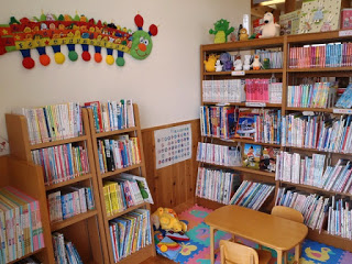カラフルなプレイマットが敷かれ、小さなテーブルと椅子が置かれたスペースの周りに、たくさんの本が並んだ本棚が複数置かれた、小さい子ども向けのコーナーの写真