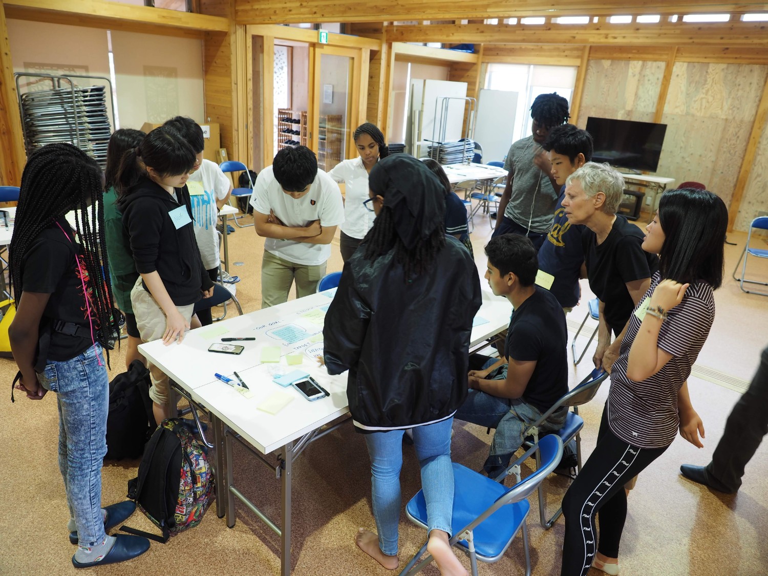 教室のような木造の建物の中で、10人以上の若者が立って一つのテーブルを囲み、アイデアが記載された付箋を真剣に見たり話し合ったりしている写真