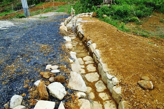 黒い砂利と茶色い土の間に、大きな石を並べて作られた排水溝のような石組みの道が写っている写真