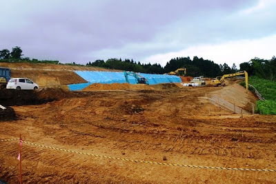 傾斜地にブルーシートが敷かれ、重機で土を掘り起こす作業が進む造成地の様子を写した写真