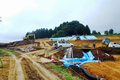 重機や作業車、配管材などが並び、工事が進行中の広い土の斜面が見える造成地の写真