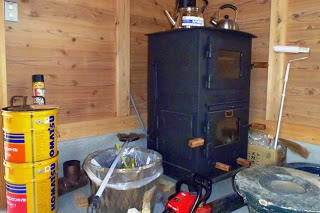 黒い薪ストーブの上に2つのやかん、周りに燃料缶、斧などが置かれた、木造の作業小屋の一角を写した写真