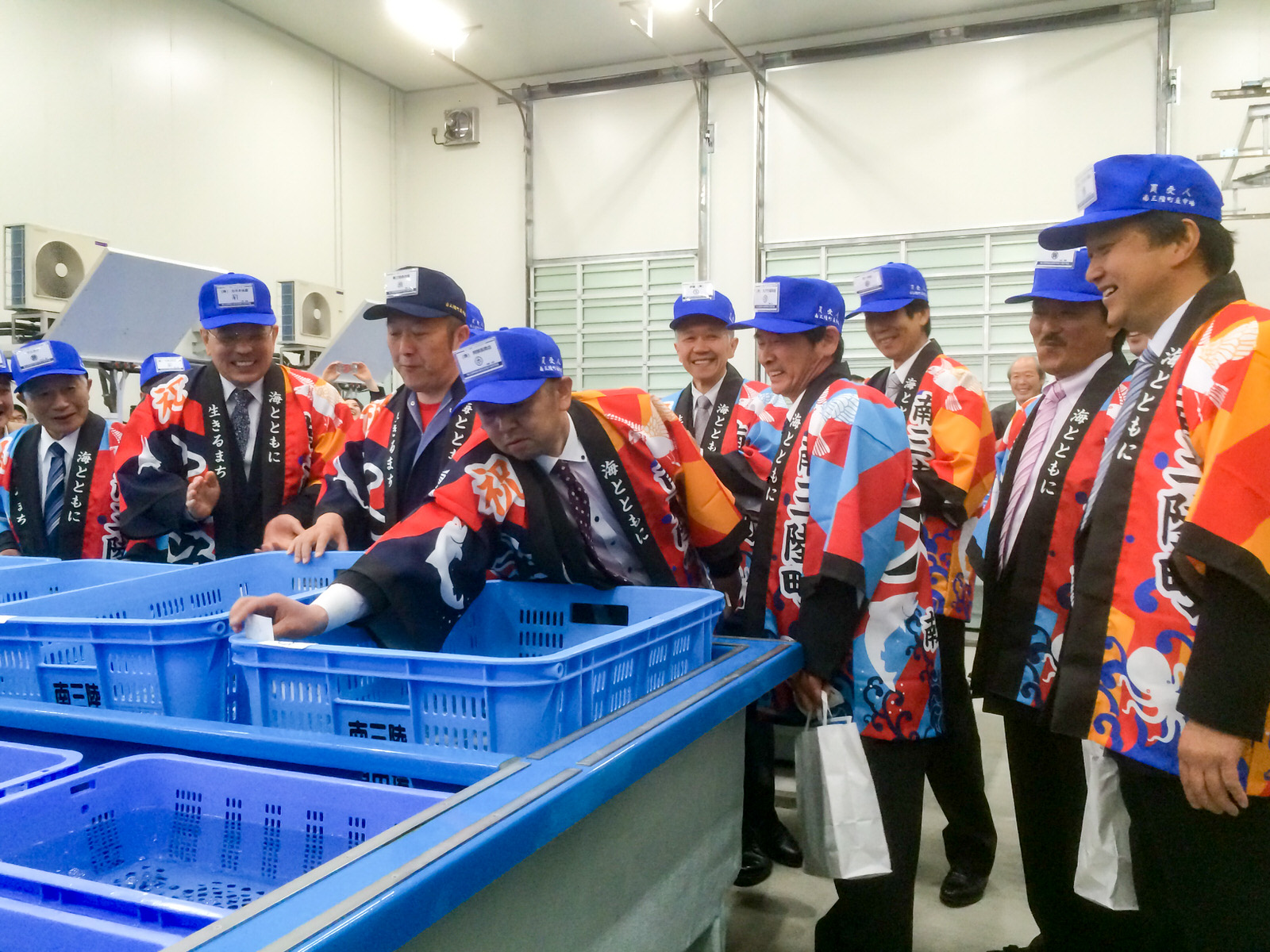 カラフルな法被と青い帽子を着た関係者たちが、水色のかごが並んだ水槽の周りに集まり笑顔を浮かべている写真