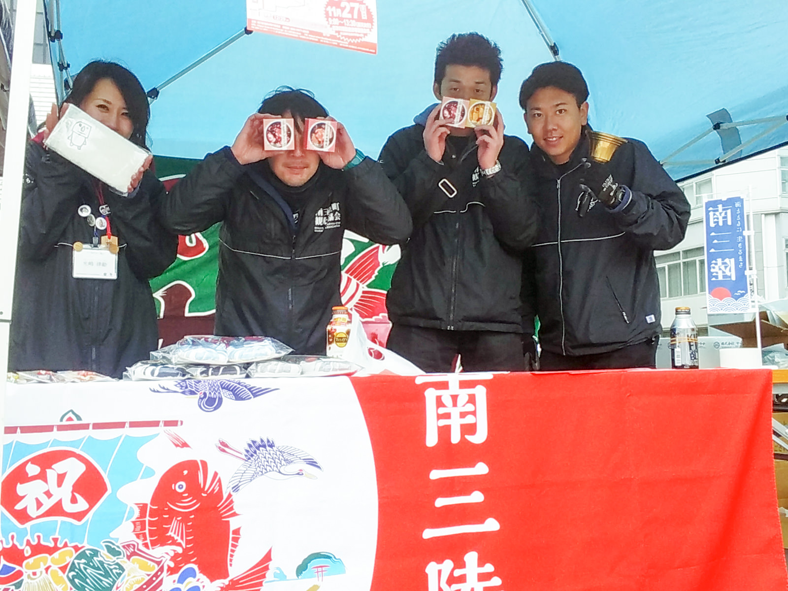 赤い大漁旗をかけたテーブルの後ろで四人のスタッフが並び、商品やベルを手にして笑顔でポーズを取っている様子の写真