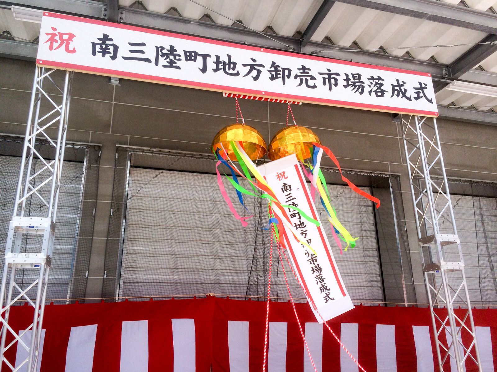 「祝 南三陸町地方卸売市場落成式」と書かれた横断幕が掲げられたゲートの中央に吊るされた金色のくす玉が割られ、「祝 南三陸町地方卸売市場落成式」の垂れ幕とカラフルなテープが風になびいている様子の写真