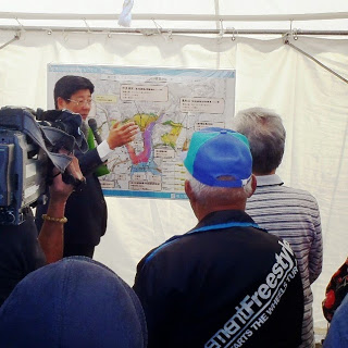 スーツ姿の男性が白いテントの中に掲示された大きな地図の前で集まった人々に説明しており、その様子をテレビカメラが撮影している写真