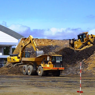 茶色い土の山を背景に、大型ダンプとショベルカーが作業中で、ブルドーザーも稼働している写真