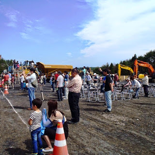 開けた敷地に重機とパイプ椅子が並び、家族連れなどの来場者が集まって見学している様子の写真
