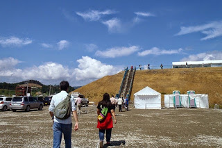 仮設トイレと白テントが並ぶ広場を歩く人々の先に、長い階段が設置されている土でできた高台の写真