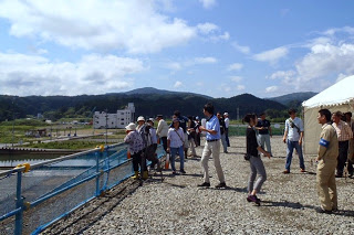 天気の良い青空の下、右側にテントが設置された傍で女性が左腕に腕章をつけたスタッフと会話している様子や、高台の上から柵越しに町の様子を見学する人々の写真