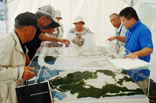 白いテントの中で、青いシャツを着た担当者が透明カバーの上から地形の立体模型を指しながら、年配の男女数人に対して復興計画の説明をしている写真