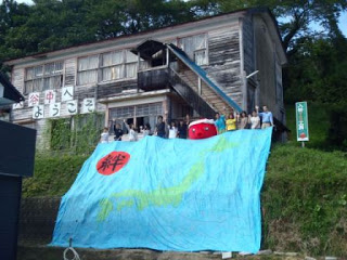 古い木造の建物の前の斜面に、日本列島の地図と赤い丸に「絆」の漢字が描かれている大きな青い布が広げられ、その上にオクトパス君の着ぐるみと複数の人々が立っている写真