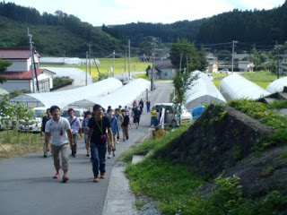 複数並んだ農業用のビニールハウスの間を通り、まっすぐのびた道を、多数の参加者が集団で歩き、地域の農地や復興の現場を見学している様子の写真
