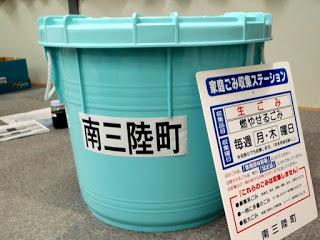 「南三陸町」と書かれた水色の大型プラスチックバケツが床の上に置かれていて、横にはごみ収集日の案内板が立てかけられている写真