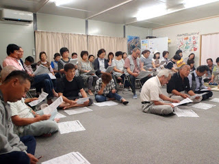 明るい室内で地域住民が床や椅子に座って配布資料を見ながら説明を真剣に聞いている住民説明会の様子の写真