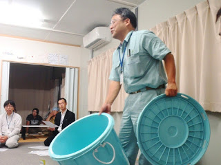 作業服を着た男性が水色のバケツの本体とふたを手に持ち、参加者に見えるように説明をしている写真