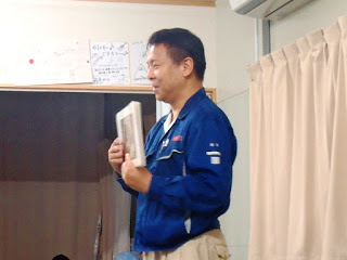 青いジャンパーを着た男性が立って紙芝居風のパネルを持ち話をしている写真