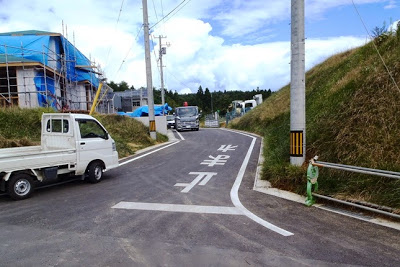 「止まれ」と書かれた細い舗装道路上に軽トラックや大型トラックが停まっており、左側にビニールシートに覆われた建設中の住宅がある写真