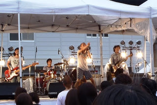 屋外の特設ステージで5人編成のロックバンドが演奏を行い、ステージ前には観客が集まり音楽を楽しんでいる様子の写真