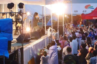 ライトに照らされた夜の屋外ステージで歌手が情熱的に歌い、その下でたくさんの観客たちが手を挙げて演奏を楽しんでいる様子の写真