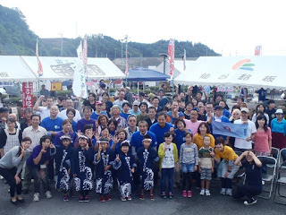 イベント会場のテント前に大人から子どもまで多くの参加者が集まり、笑顔でカメラに向かってポーズをとっている集合写真