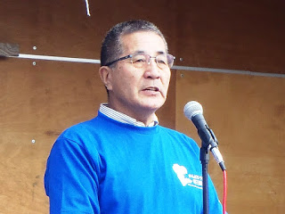 青い長袖シャツを着た男性がマイクを前に立ち、話をしている様子の写真