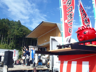 青空の下、山を背景にした屋外ステージで数人が手を挙げている様子と、前景に赤いタコのオクトパス君の乗った神輿の飾りや「タコまつり」と書かれた旗が並ぶ写真