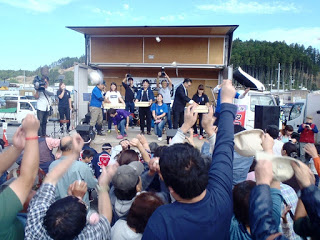 屋外ステージ上から餅などが投げられ、集まった多くの人々が手を伸ばして受け取ろうとしている賑やかな様子の写真