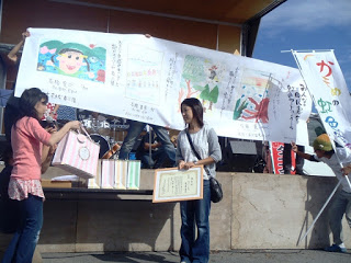 屋外ステージで大きな絵やメッセージが描かれた作品が数点並んだ長い模造紙を掲げ、その前で女性が表彰状を持った女性に景品を手渡す様子が写された写真