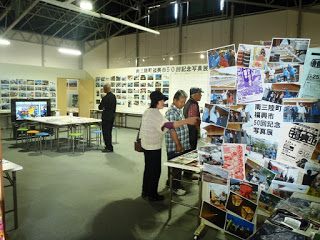 屋内の展示会場で、壁やパネルに多数の写真が掲示され、来場者が写真展を鑑賞している様子の写真