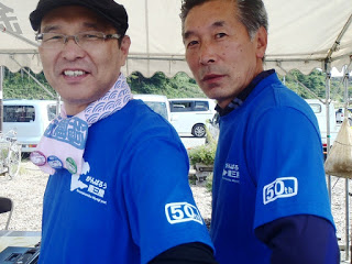 屋外テントの下で青い「50th」のロゴ入りTシャツを着た二人の男性が並び、カメラに向かって微笑む様子の写真