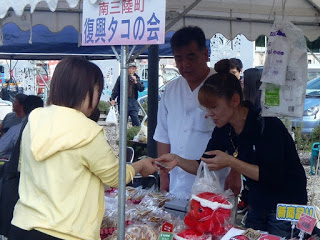 「南三陸町復興タコの会」と書かれたテントの下で、女性店員が来場者に商品を手渡し、男性店員が見守る屋台販売の様子を撮影した写真