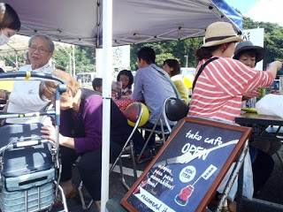 屋外テントの下に並べられた椅子とテーブルで人々が飲食を楽しみ、前景に「tako cafe OPEN」と書かれた看板が立つ様子を撮影した写真