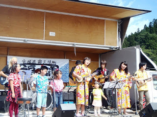 屋外ステージで色鮮やかな衣装を着た女性や子どもたちが楽器を演奏し歌い、後方でバンドメンバーが伴奏する様子の写真