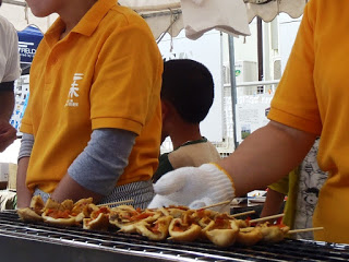 黄色いシャツを着た屋台のスタッフが、焼き網に串を並べて調理している様子の写真