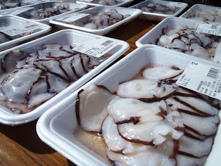 テーブルに並べられた発泡トレイ入りのスライスされた生タコが、ラベル付きで販売されている様子の写真