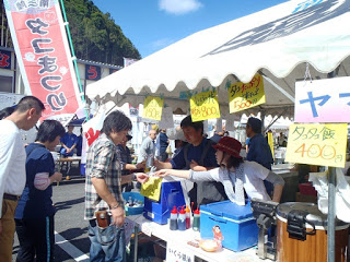 「タコ唐揚げ」や「タコ飯」などの手書き看板を掲げた屋台で、店員が来場者に商品を手渡している福興市の様子を撮影した写真
