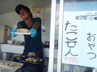 屋台の店員がたこ焼きをせんべいで挟んだ「たこせん」を笑顔で差し出し、横には手書きで「おやつ たこせん 300円」と書かれた紙が貼られている写真