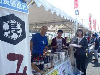 白いテントの下、醤油瓶や鍋が並ぶ屋台で男性店員二人が立ち、女性客が商品を手に笑顔を見せている様子の写真