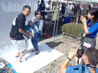 屋外で、白い布の背景の前に立つ赤ちゃんを抱いた男女がカメラマンに撮影され、横で女性スタッフが笑顔で見守る様子の写真