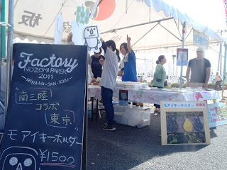 白いテントの前に「南三陸コラボ東京 モアイキーホルダー ¥1500」と書かれた黒板が立ち、奥でスタッフが接客や作業をしている様子の写真