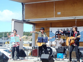 青空の下、屋外ステージで4人編成のバンドがギターやドラムを演奏しながら歌い、観客の方を指さしている様子の写真