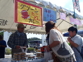 白いテントの下、「グリーンたこさんカレー」や「海鮮チヂミ」と書かれた看板が掲げられ、男性店員が来場者にパック詰めの商品を販売している様子の写真